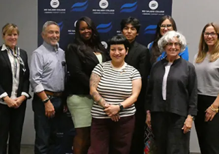 A group of eight adults stand together smiling in front of a branded backdrop, posing for a photo at a recognition event.