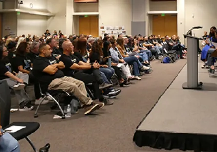 A large audience seated in a conference room facing a stage where speakers are presenting during an event.