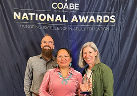 Three people stand smiling in front of a backdrop reading “COABE National Awards Honoring Excellence in Adult Education,” with one person holding an award.