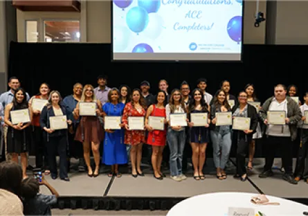 Large group of ACE program completers and staff pose on stage holding certificates, smiling in front of a screen that reads “Congratulations ACE Completers.”