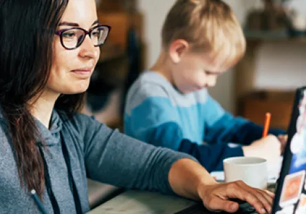 Adult learner working on a laptop at home while helping a child with schoolwork, illustrating flexible online learning in a real-life setting.