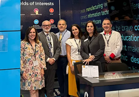 **Alt text:** Six professionals stand together smiling at a conference exhibit booth during an education and workforce event.