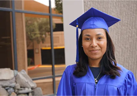 Portrait of Ebony Livas wearing a blue graduation cap and gown, standing outdoors and looking at the camera.
