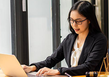 Woman wearing glasses and business attire works on a laptop at a desk, with a gavel and scales of justice nearby.