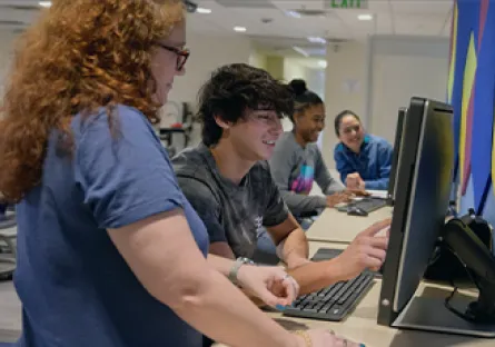 An instructor stands beside a student as they work together at a desktop computer in a classroom, while three other students use computers in the background, illustrating collaborative, technology-enabled learning.