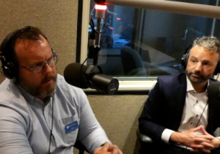 Ken Wilk and Zach Mannheimer wearing headphones speak into microphones during a podcast recording in a radio studio.