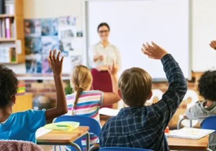 Students raise their hands to participate in a classroom discussion while a teacher stands at the front of the room.