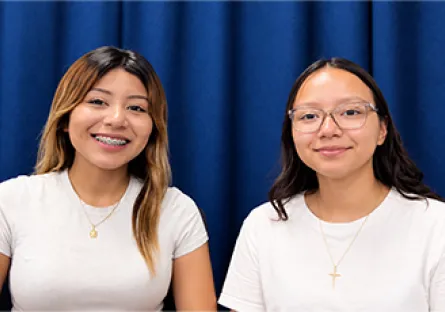 Allyson Hurtado Fernandez, left, and Silvana Castro Aguilar, right, smile at the camera while seated together in front of a blue curtain.