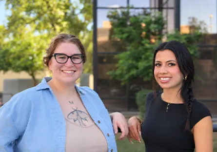 Maren “Rain” Colon, left, and Ariel Davila, right, smile together outdoors in front of a glass building and greenery.