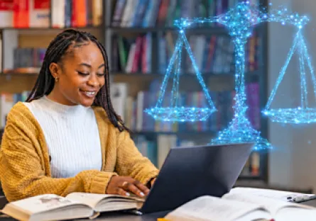 A smiling woman with long braids wearing a mustard-yellow cardigan works on a laptop at a library table with open textbooks. A glowing blue digital illustration of the scales of justice appears above the laptop, representing legal studies and online education.