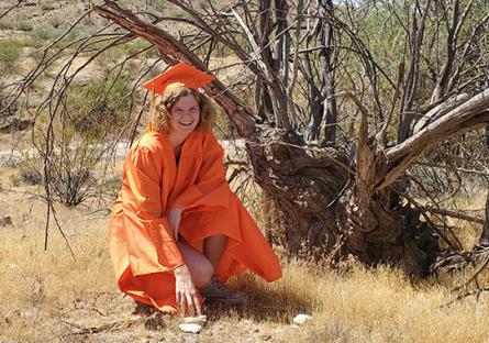 photo of Emma Harlow in a desert landscape kneeling under a dead tree in her graduation regalia.